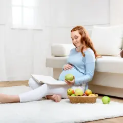 Kobieta w ciąży czyta książkę, trzymając jabłko. Obok koszyk z owocami. Dba o dietę, by uniknąć listeriozy w ciąży.