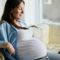Kobieta w zaawansowanej ciąży, z widocznym brzuszkiem, siedzi na kanapie. To piękny czas stawiania się macicy i oczekiwania na dziecko.