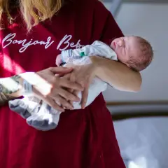 Sennik poród: noworodek śpi w ramionach osoby z tatuażem na ramieniu, ubranej w bordową koszulkę z napisem "Bonjour Baby".