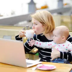 Kobieta pije kawę, trzymając dziecko. Obok leży laptop. Karmienie piersią i chwila dla siebie.