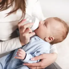 Mama karmi noworodka butelką. Delikatne dłonie trzymają smoczek, a maluch z zapałem pije mleko.