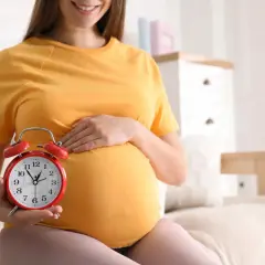 Uśmiechnięta ciężarna kobieta trzyma czerwony budzik, symbolizując zbliżający się poród indukowany.