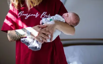 Sennik poród: noworodek śpi w ramionach osoby z tatuażem na ramieniu, ubranej w bordową koszulkę z napisem "Bonjour Baby".