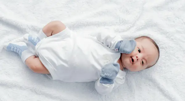 Malutki noworodek w białym body i niebieskich skarpetkach, z rękawiczkami na rączkach. Wygląda, jakby zastanawiał się, kiedy wreszcie zrobi kupę.