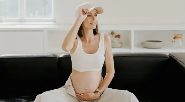 Kobieta w ciąży, w białym topie i czapce, siedzi na czarnej kanapie, czując uderzenia gorąca.