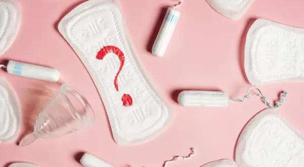 Różne produkty higieniczne, w tym podpaski, tampony i kubeczek menstruacyjny, ułożone na różowym tle. Na jednej z podpasek widnieje czerwony znak zapytania, symbolizujący brązowe plamienie.