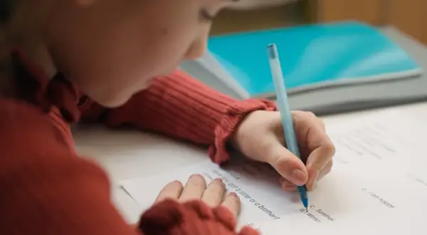 Dziecko w czerwonym swetrze pisze długopisem po kartce. Czy to zadanie z języka angielskiego, od jakiej średniej jest 5?