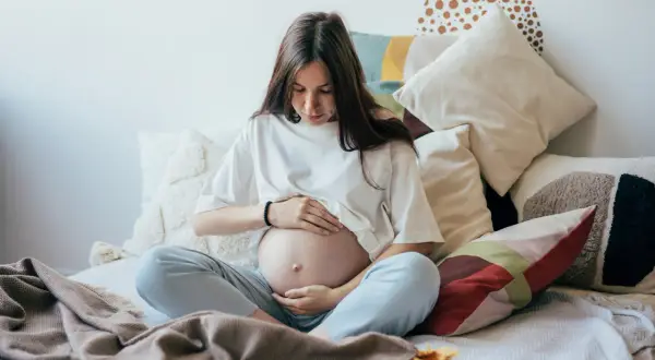 Kobieta w ciąży, otoczona poduszkami, czule dotyka brzucha. Czas oczekiwania na dziecko, być może zmagająca się z wielowodziem, ale pełna spokoju.