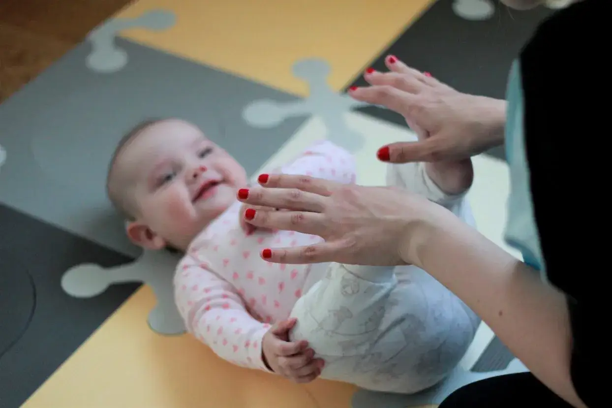 Uśmiechnięte 5 miesięczne dziecko leży na macie, a dorosły bawi się z nim, dotykając jego nóżek.