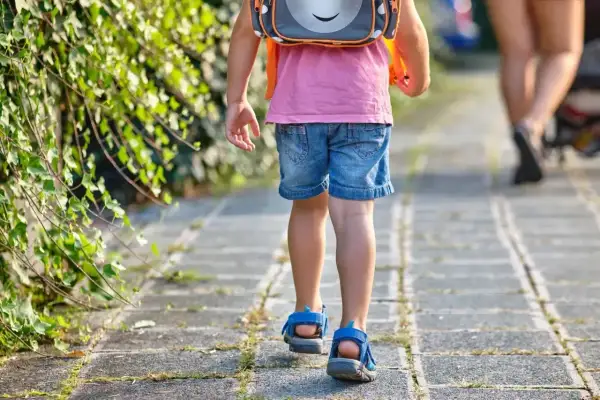 Małe dziecko w różowej koszulce i dżinsowych szortach z plecakiem idzie chodnikiem. To widok, kiedy dzieci idą do szkoły.